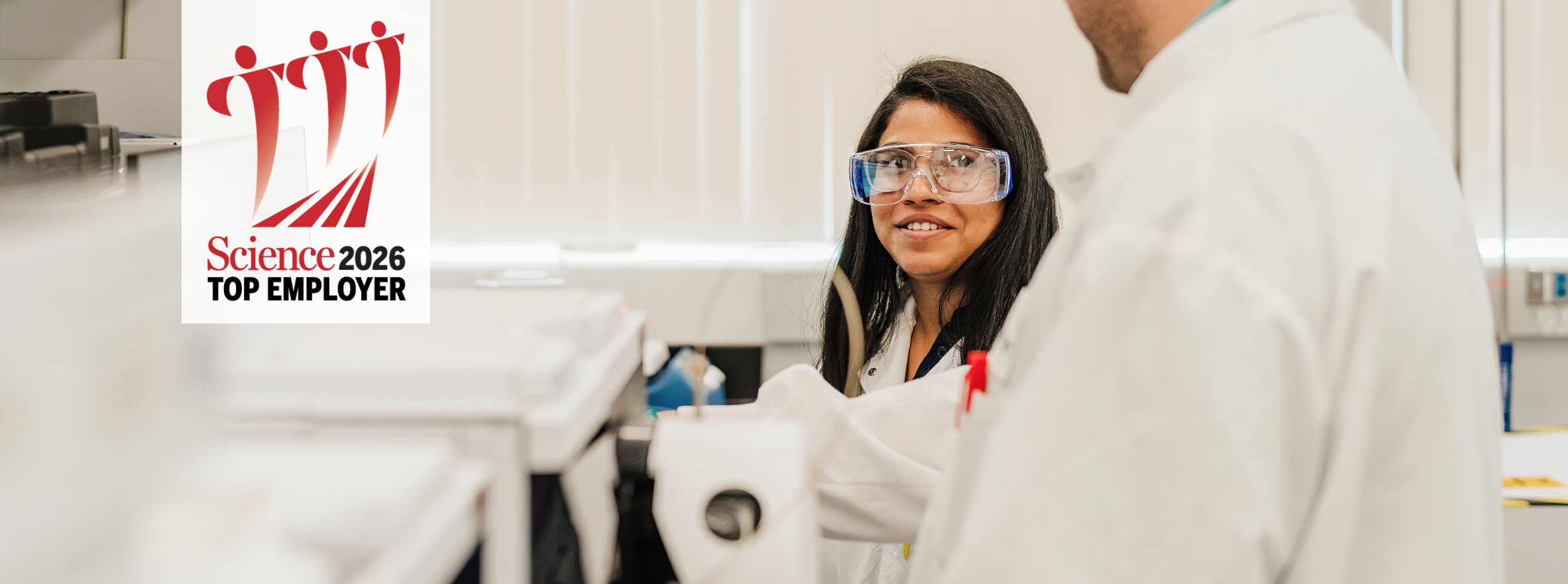 A female scientist wearing safety glasses and a lab coat in a lab talking with a colleague