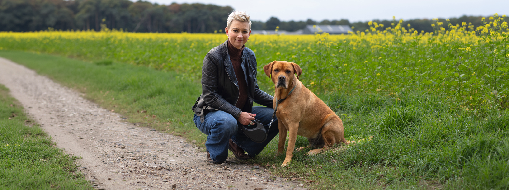 Ester and her dog kneel on the side of a country path with green fields stretching into the distance