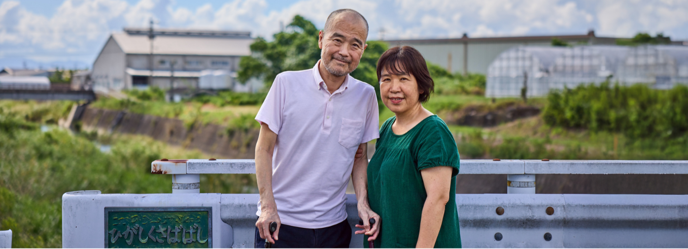 A Japanese patient and his wife stand on a bridge looking at the camera with fields and greenhouses behind them